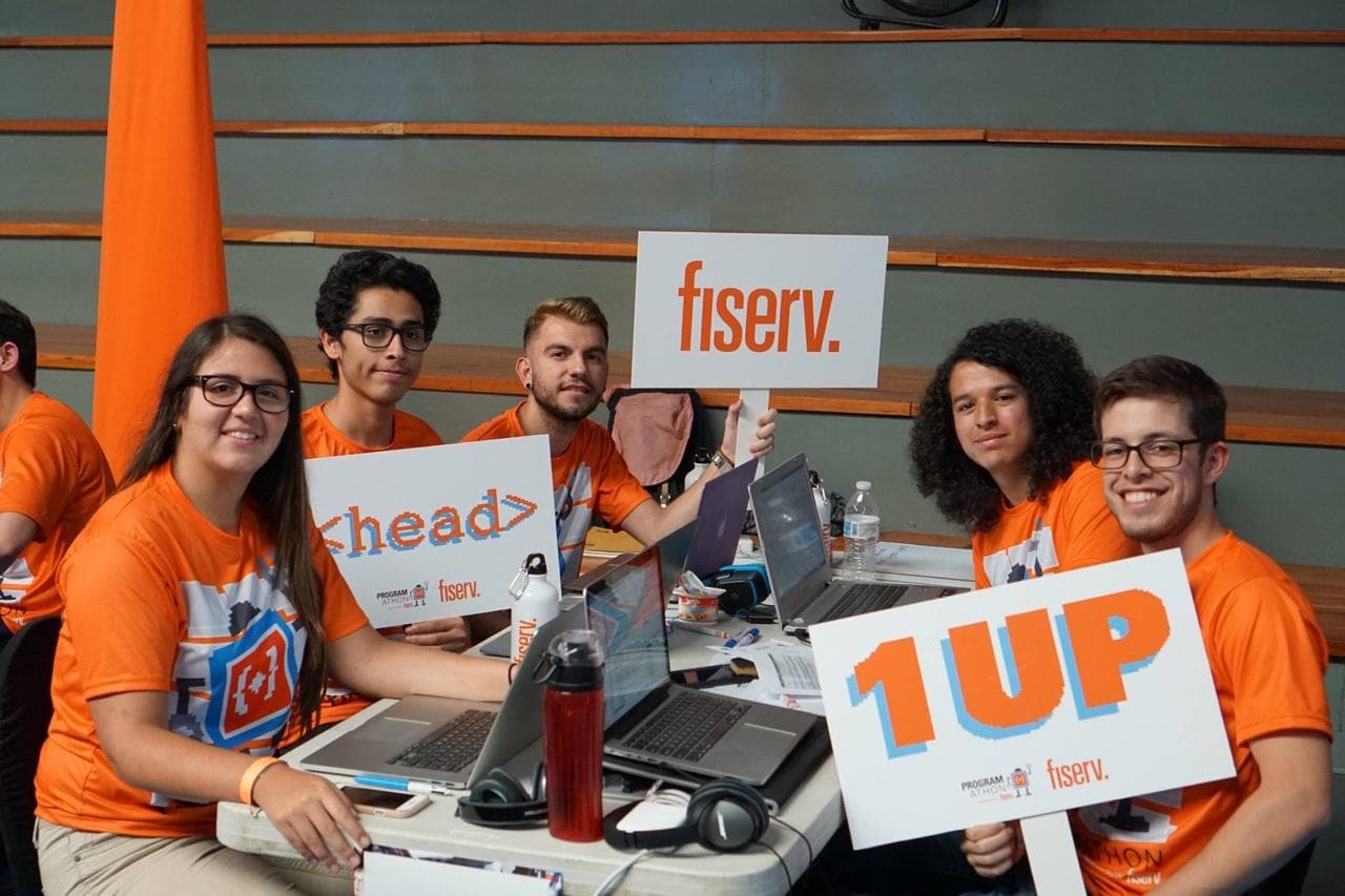 Team String Cheese in orange Programathon shirts holding Fiserv, <head> and 1UP signs